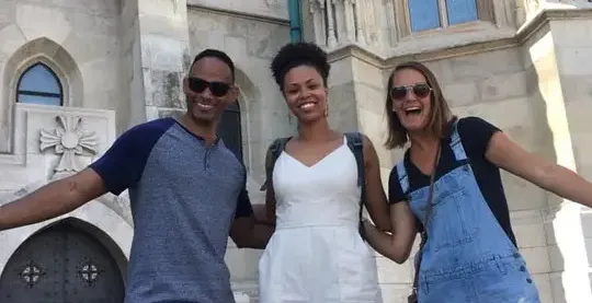 Three smiling people stand arm-in-arm in front of the ornate, light-colored Matthias Church with Gothic windows.