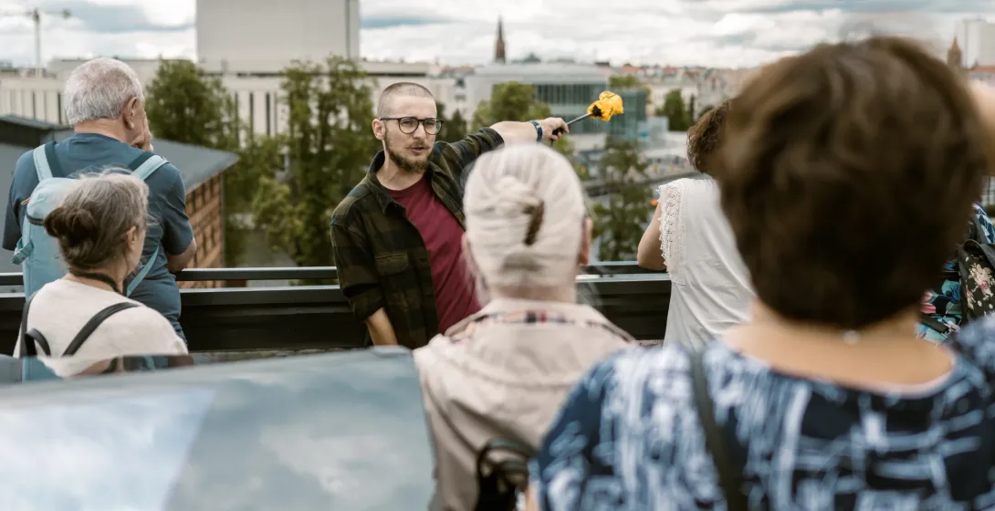 A tour guide with glasses points across the Bydgoszcz city skyline while explaining something to a small group of tourists.