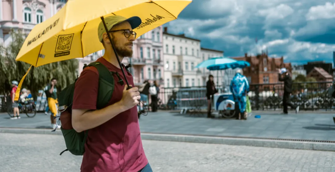 Przewodnik z brodą i w okularach trzyma duży żółty parasol Walkative, prowadząc wycieczkę po europejskim mieście.