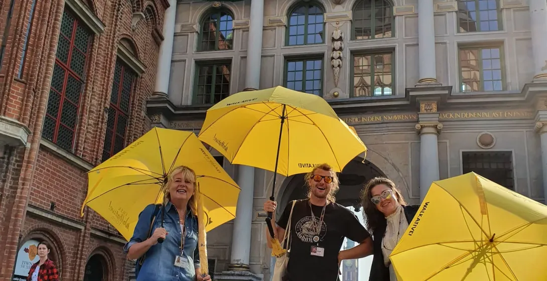 Tres guías sonrientes de Walkative con paraguas amarillos en una calle empedrada frente a la histórica Puerta Verde de Gdańsk