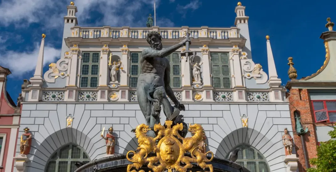 Bronzestatue des Neptun mit Dreizack auf einem Brunnen vor einem prächtigen, reich verzierten Gebäude bei blauem Himmel.