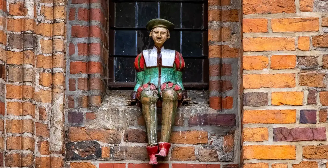 Bunte Holzfigur eines Mannes mit grüner Baskenmütze und mittelalterlicher Kleidung, sitzend auf einer Ziegel-Fensterbank e...