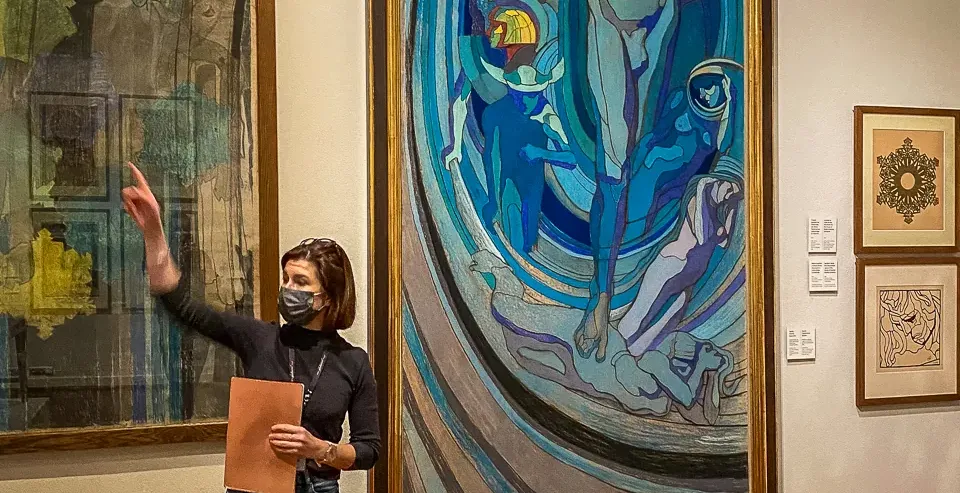 Eine Stadtführerin mit Gesichtsmaske steht in einer Kunstgalerie und zeigt auf ein großes, farbenfrohes Gemälde an der Wand.