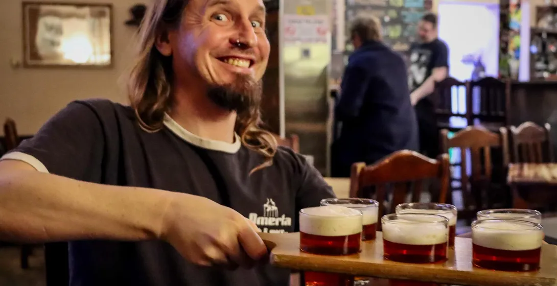 Man with long hair and beard holding a beer flight with five small glasses of amber beer in a pub.