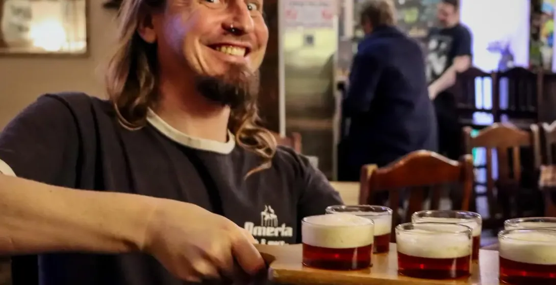 Man with long hair and beard holding a beer flight with five small glasses of amber beer in a pub.