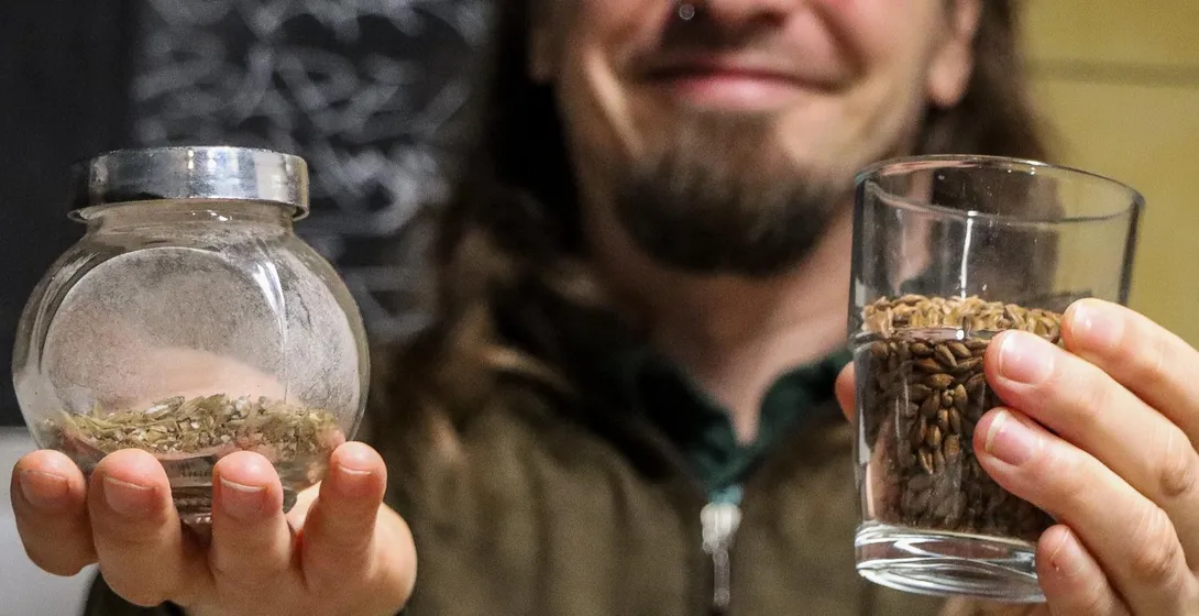 Ein Mann mit langen Haaren hält ein Glas mit Gerstenmalz und ein kleines Glas mit getrocknetem Hopfen in die Kamera.