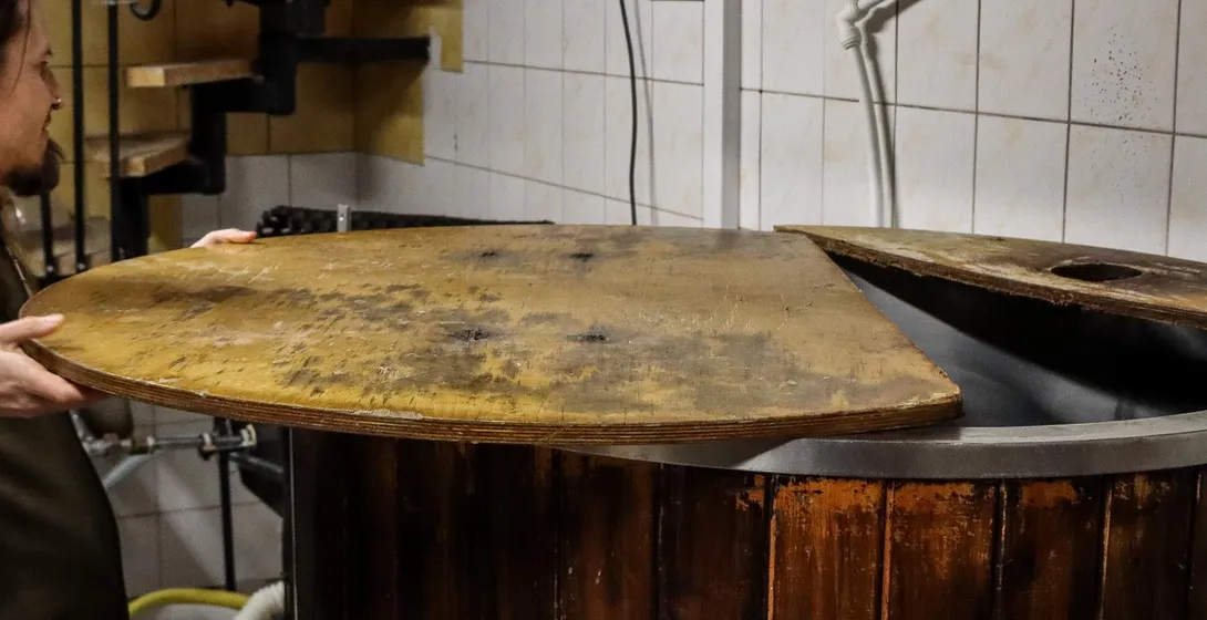 Ein Brauer öffnet den Holzdeckel eines großen Gärbottichs in einer kleinen Brauerei.