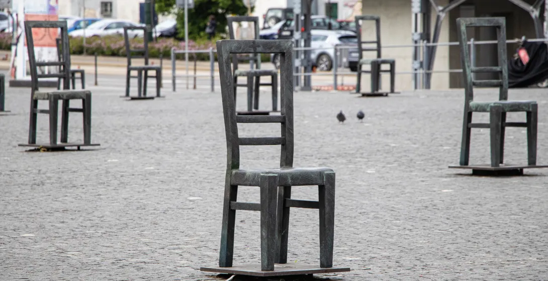 Esculturas de sillas de metal llenan una plaza adoquinada, con dos palomas visibles y edificios al fondo.