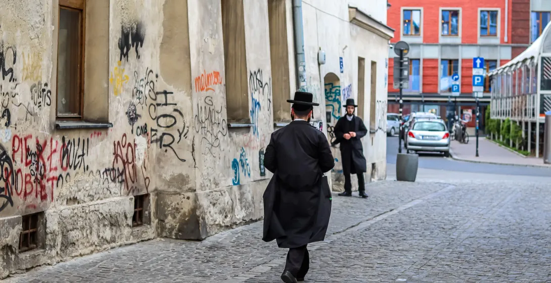 Two Orthodox Jewish men on a cobblestone street in Krakow's Kazimierz district, near a wall covered in graffiti.