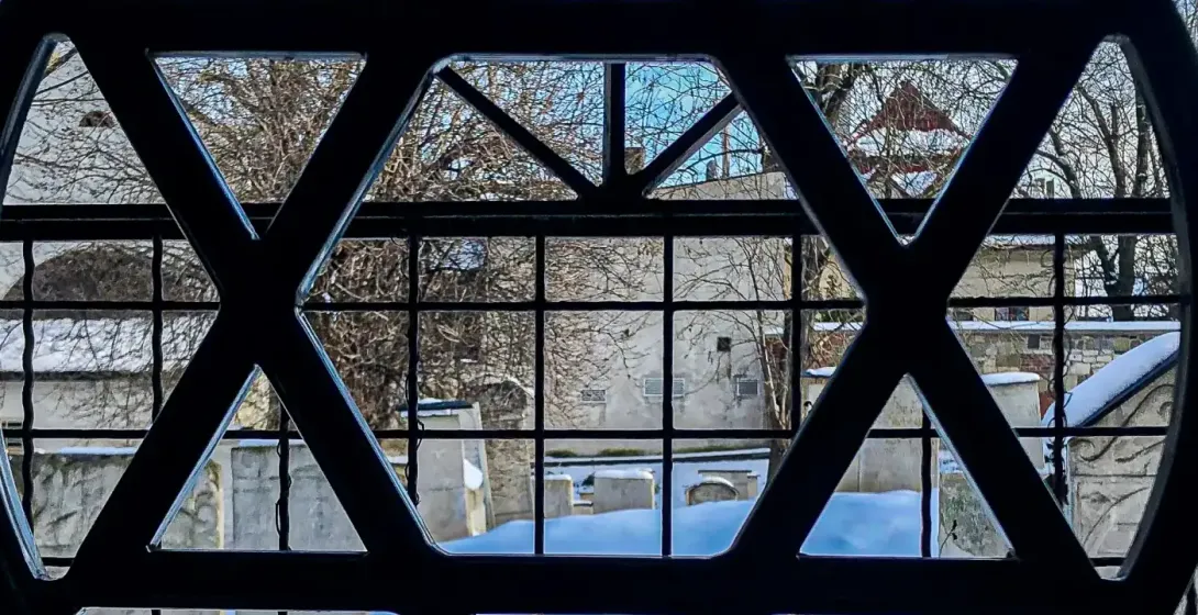 Blick durch ein Fenster mit einem Davidstern-Gitter auf einen schneebedeckten Friedhof im Winter.