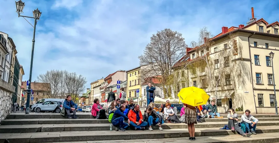 Eine Reisegruppe sitzt auf breiten Steinstufen auf einem Stadtplatz in Krakau und lauscht einem Führer mit gelbem Schirm.