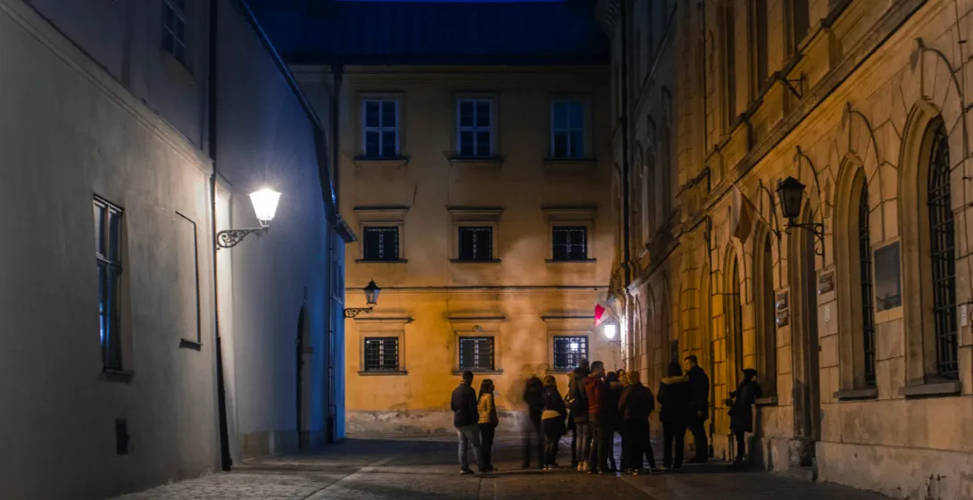 A tour group stands on a narrow, lamplit cobblestone street in Krakow at night, surrounded by historic buildings.