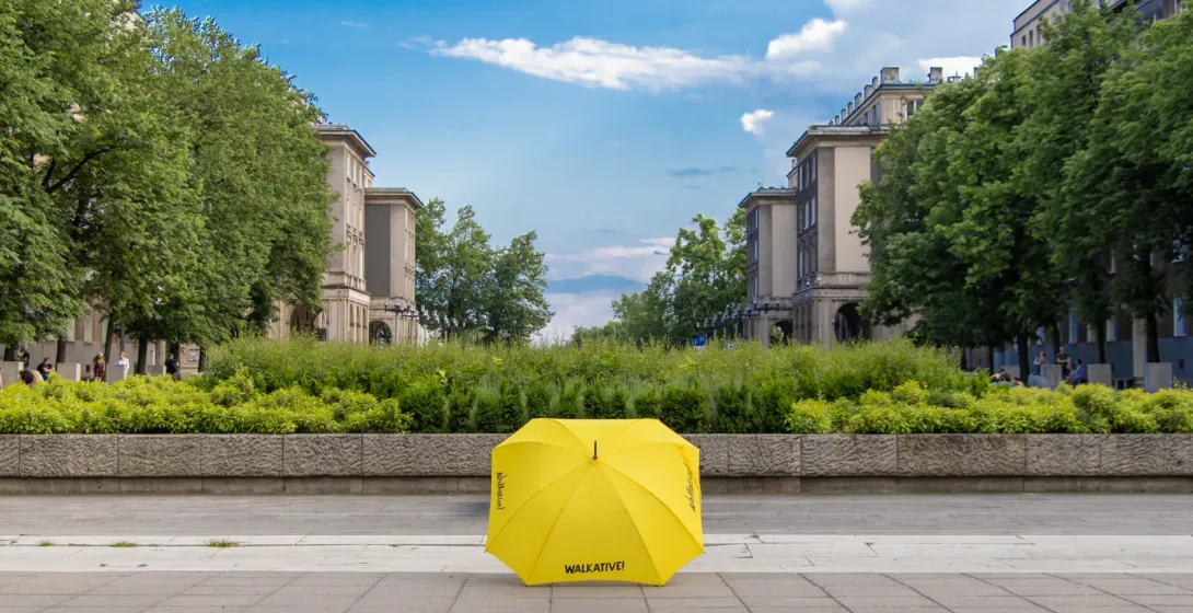 Ein gelber Walkative-Regenschirm auf dem Gehweg eines großen Platzes in Nowa Huta, mit Bäumen und sozialistischen Gebäuden.