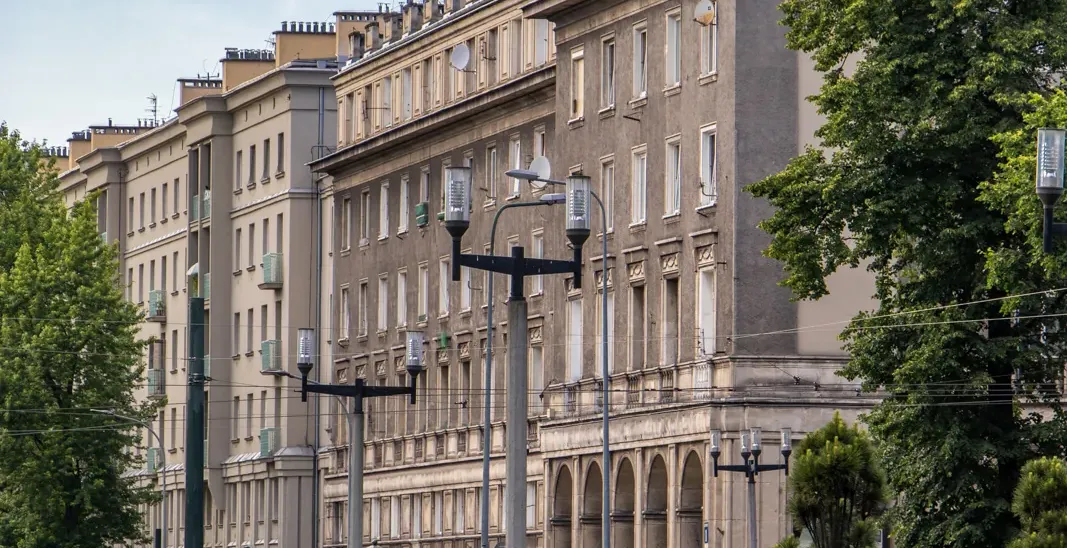 A wide street in Nowa Huta, Krakow, with long socialist realist apartment blocks, green trees, and a blue sky.