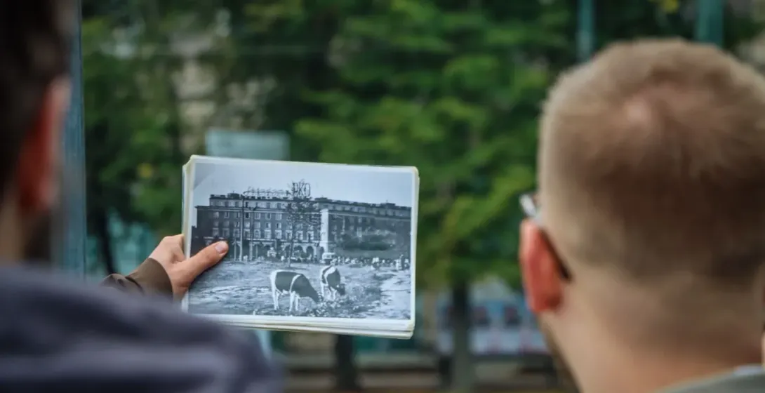A tour guide shows a visitor a historical black and white photo of a building with cows grazing in front of it in Nowa Huta.