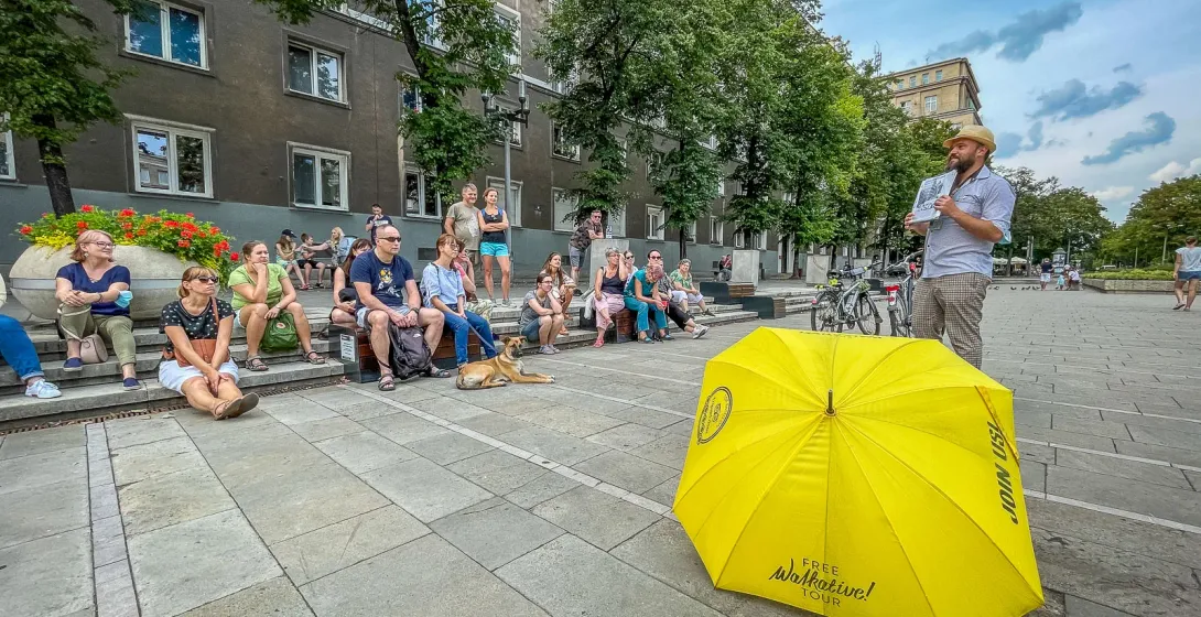 A tour guide speaks to a group of people sitting on steps in a city square, with a yellow Walkative umbrella in front.