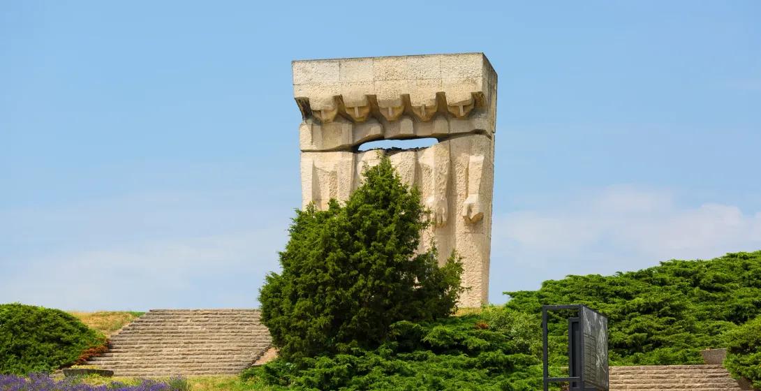 Ein großes, abstraktes steinernes Denkmal steht vor einem klaren blauen Himmel, umgeben von grünen Sträuchern, Steintreppe...
