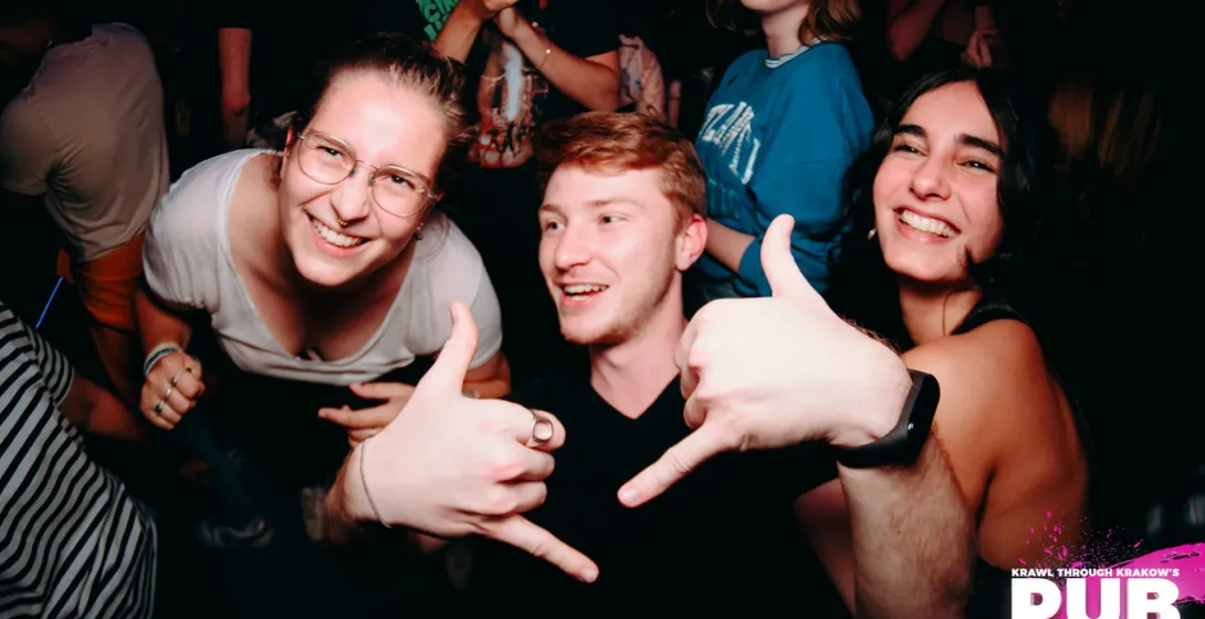 Grupa młodych ludzi uśmiecha się i pozuje do zdjęcia w zatłoczonym, słabo oświetlonym klubie lub barze.