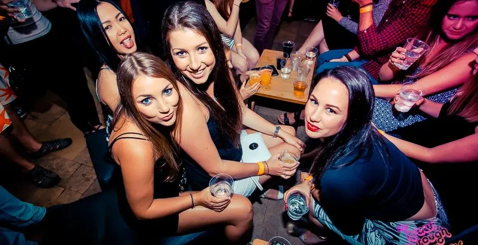 Grupa młodych ludzi, głównie kobiet, towarzysko pije napoje w słabo oświetlonym barze, uśmiechając się do aparatu.