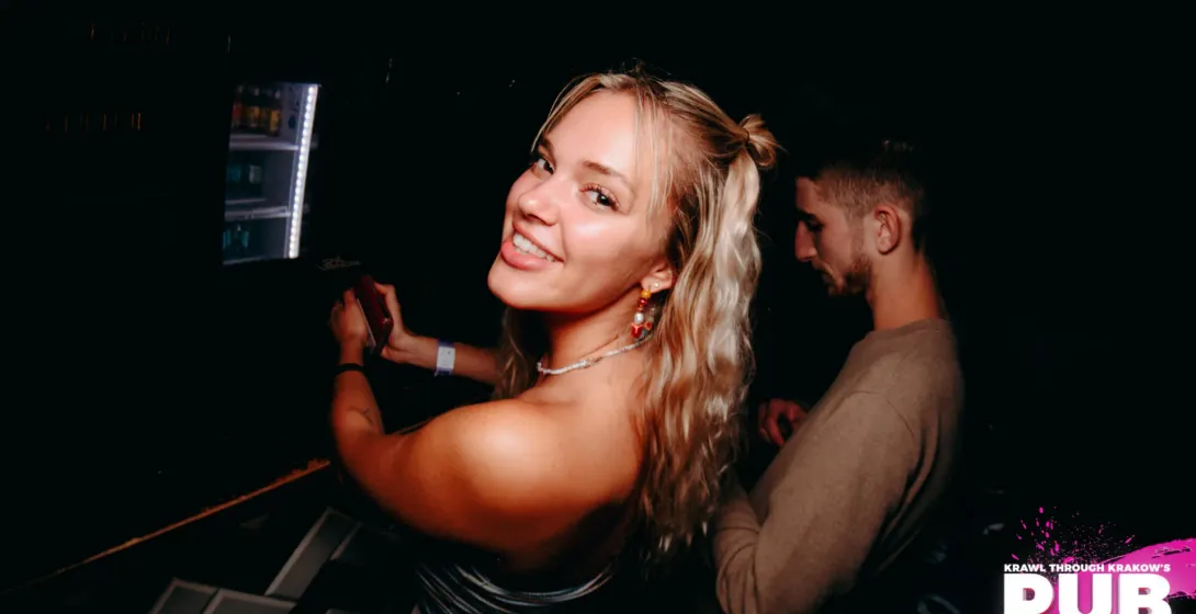 Uśmiechnięta młoda kobieta w słabo oświetlonym barze, trzymająca telefon, z inną osobą w tle. W tle widoczna witryna chłodnic