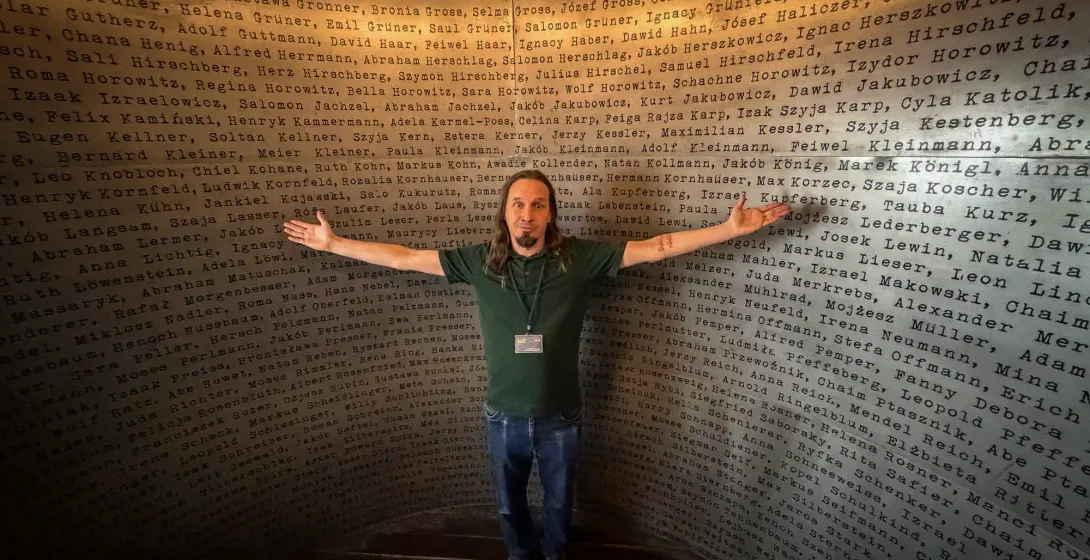 A person stands with arms outstretched in front of a wall etched with names at Schindler's Factory Museum in Krakow, creat...