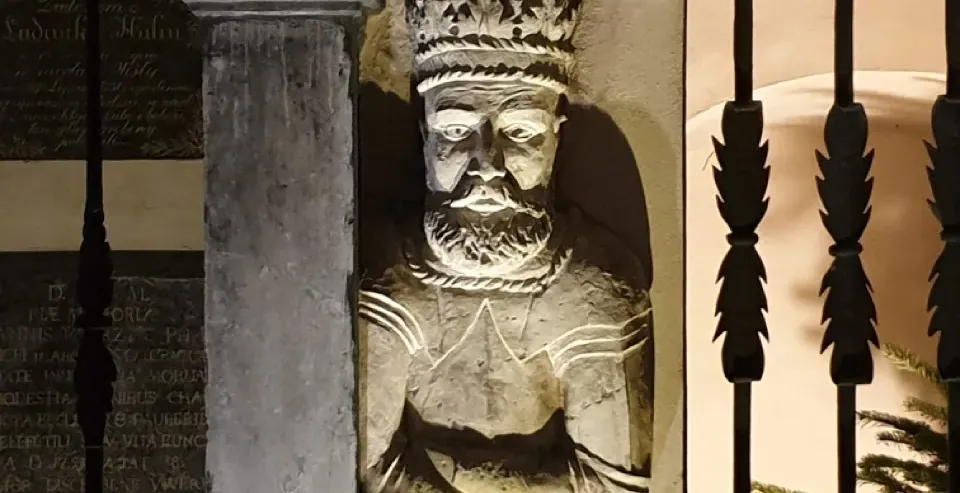 A weathered stone sculpture of a bearded king with a crown, built into a pillar and illuminated from below at night.