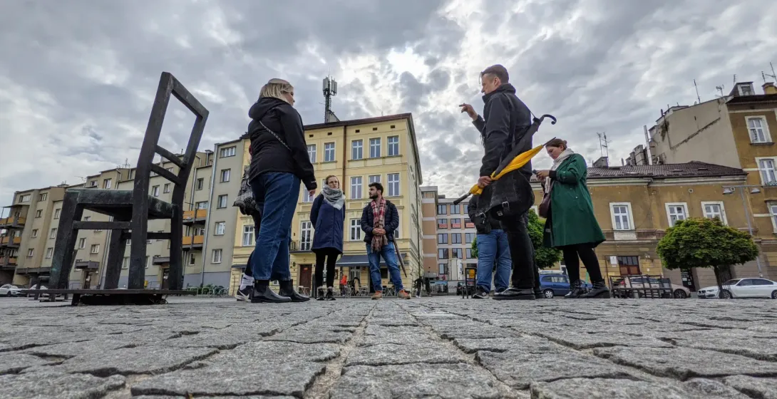 Un guía turístico habla con un pequeño grupo en una plaza adoquinada con grandes sillas de metal y edificios bajo un cielo...