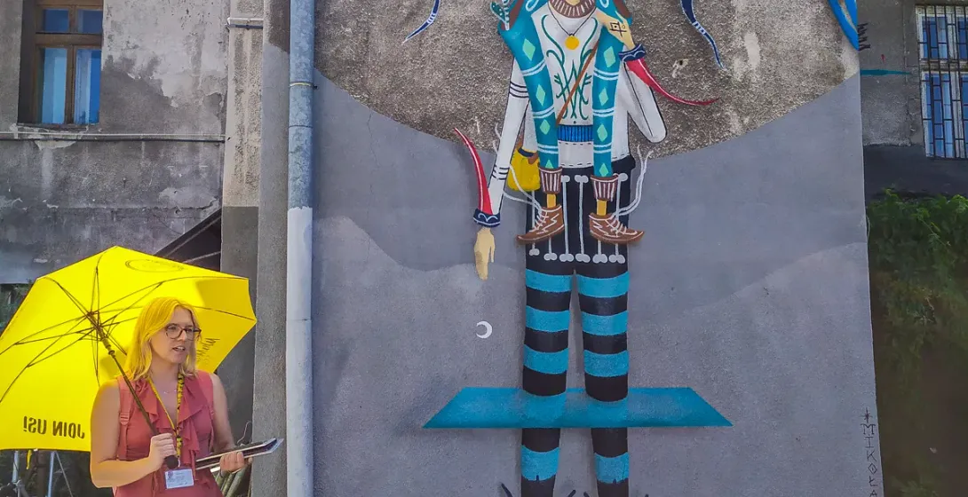 Una guía turística con un paraguas amarillo de pie frente a un gran mural colorido en un edificio de Cracovia.