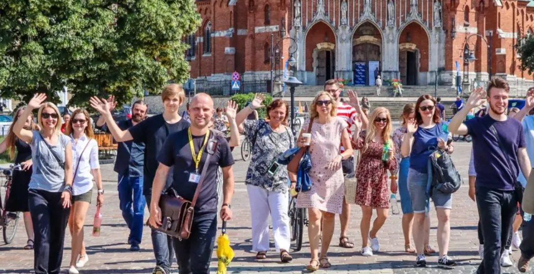 Ein Reiseführer mit gelbem Schirm führt eine Gruppe, die vor einer großen Backsteinkirche in Krakau in die Kamera winkt.