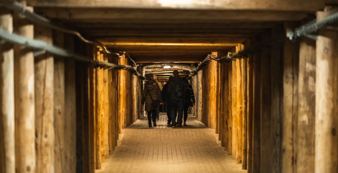 A group of tourists walking through a long, underground corridor supported by heavy wooden beams in the Wieliczka Salt Mine.