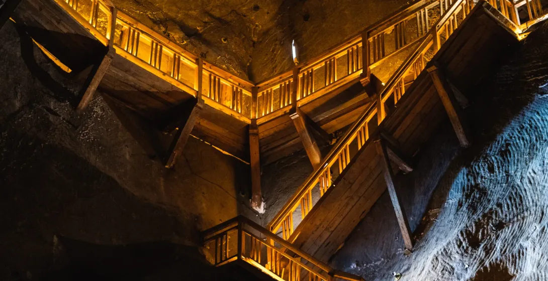 A large wooden staircase structure, lit with warm yellow light, built against the dark, rough walls of a salt mine chamber.