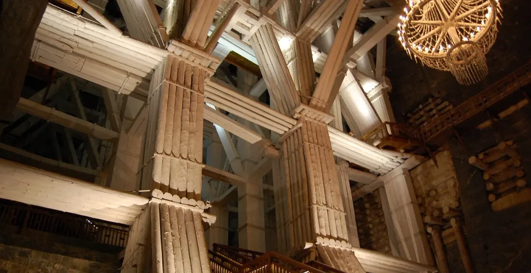 Massive wooden support beams and pillars inside the Wieliczka Salt Mine, with two large salt crystal chandeliers hanging.