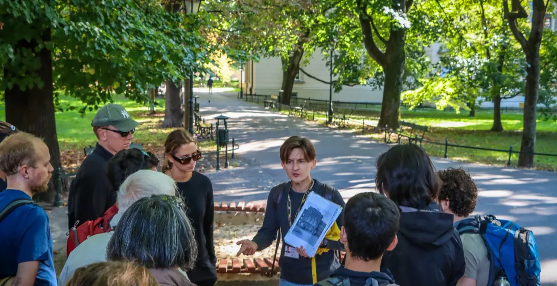 Eine Reiseführerin zeigt einer Gruppe von Personen auf einem Weg in einem sonnigen Park in Krakau historische Fotos.