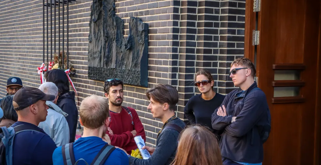 Ein Reiseführer erklärt einer Gruppe von Reisenden etwas vor einer Gedenktafel an einem Backsteingebäude in Krakau.
