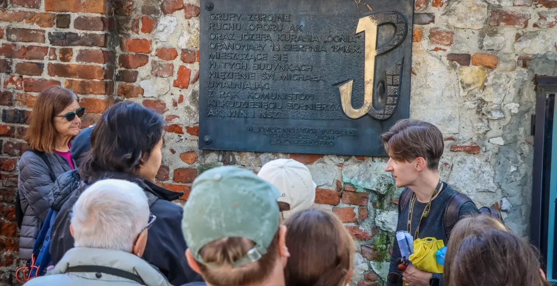 Ein Reiseführer erklärt einer Touristengruppe eine Gedenktafel mit dem Symbol des kämpfenden Polens an einer Ziegelmauer.