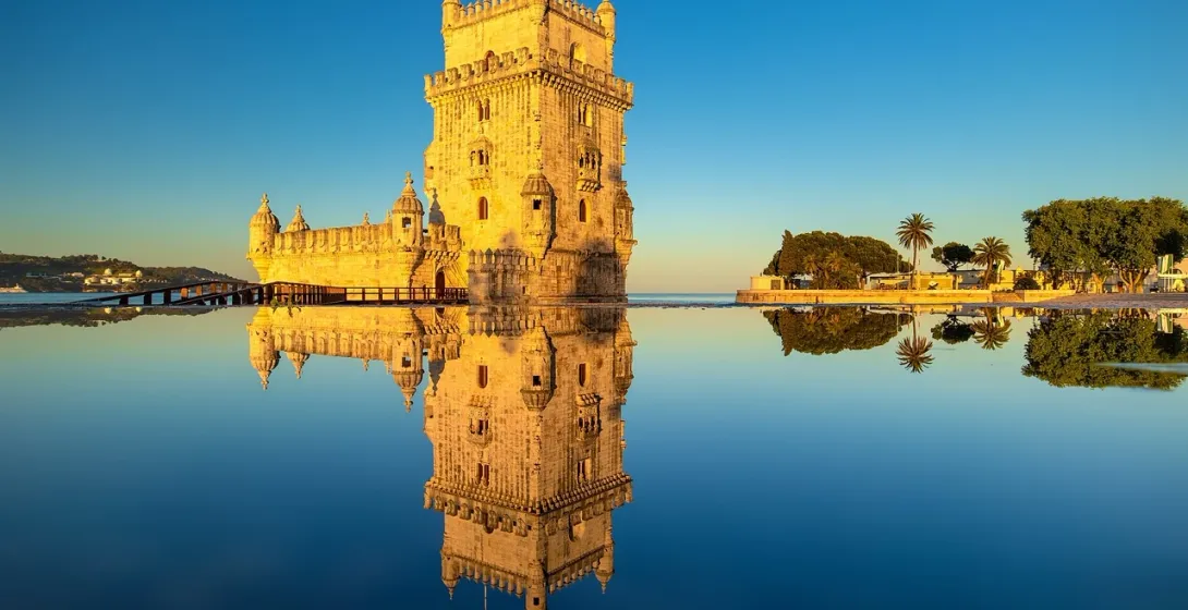 Belém Tower in Lisbon illuminated by golden light, perfectly reflected in the calm water against a clear blue sky.