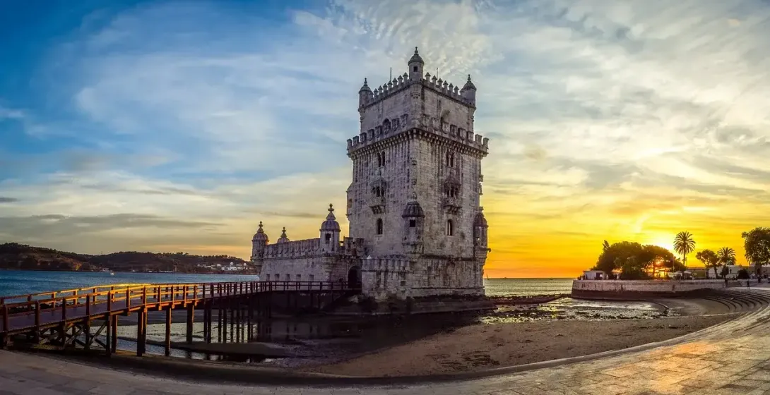 Historic Belém Tower in Lisbon at sunset, with a wooden walkway over the Tagus river and orange-blue sky.