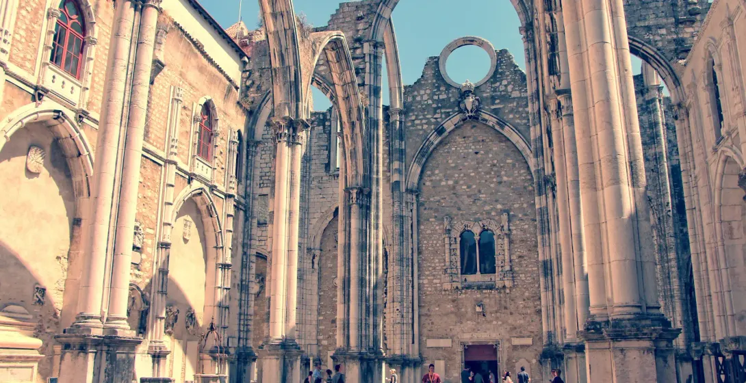 Tourists explore the roofless Gothic arches and stone ruins of the Carmo Convent in Lisbon under a clear blue sky.