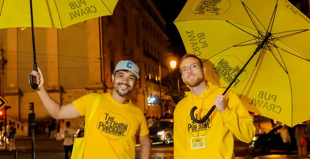 Zwei Männer in gelber 'The Lisbon Pubcrawl'-Kleidung mit gelben Regenschirmen stehen nachts auf nasser Straße in Lissabon.