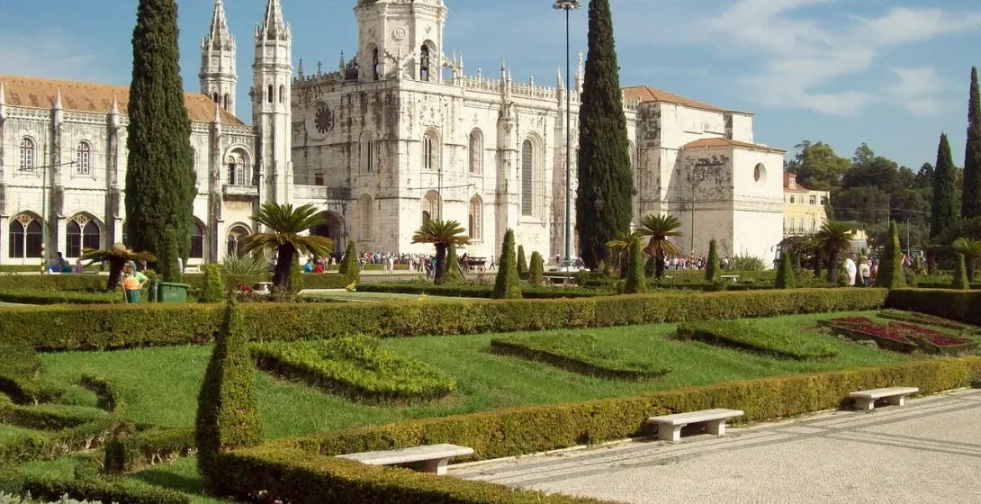 Jerónimos Monastery in Lisbon with manicured green gardens, cypress trees, and visitors on a sunny day.