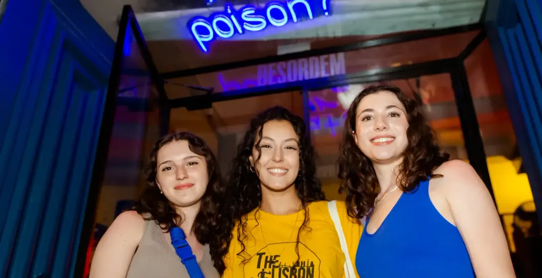 Drei junge Frauen lächeln vor einem blauen Neonschild mit der Aufschrift 'What's your poison?' über einem blauen Türrahmen.