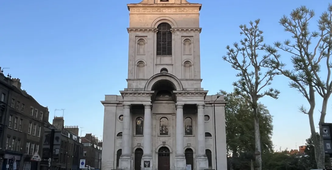 Christ Church Spitalfields seen from a cobbled street corner, its stone facade and steeple lit by the setting sun.