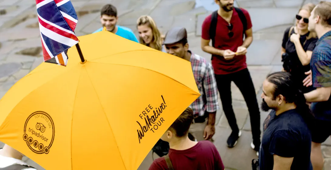 Eine vielfältige Gruppe von Menschen hört einem Reiseleiter zu, der einen gelben Walkative-Regenschirm mit kleiner Union Jack