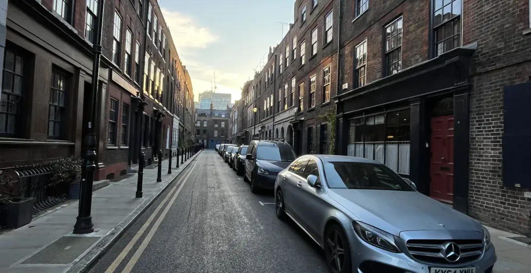 A narrow London street with cars parked between historic brick buildings at sunset.