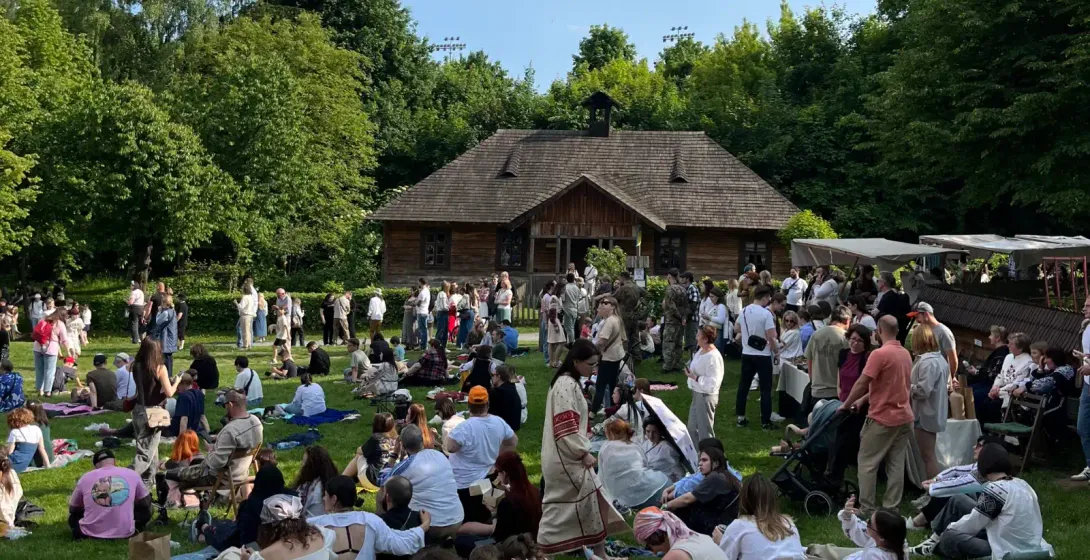 Eine große Menschenmenge versammelt sich an einem sonnigen Tag auf einer Wiese vor einem traditionellen Holzhaus.