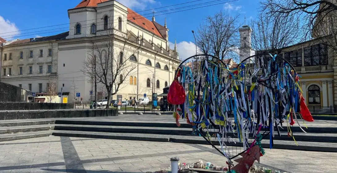 Herzförmiges Mahnmal mit blau-gelben Bändern und vielen Kerzen auf einem Platz in Lwiw, historisches Gebäude im Hintergrund.