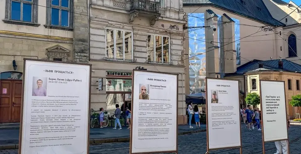 Four memorial stands with portraits and text on a cobblestone street in Lviv, with historic buildings and a church in the ...