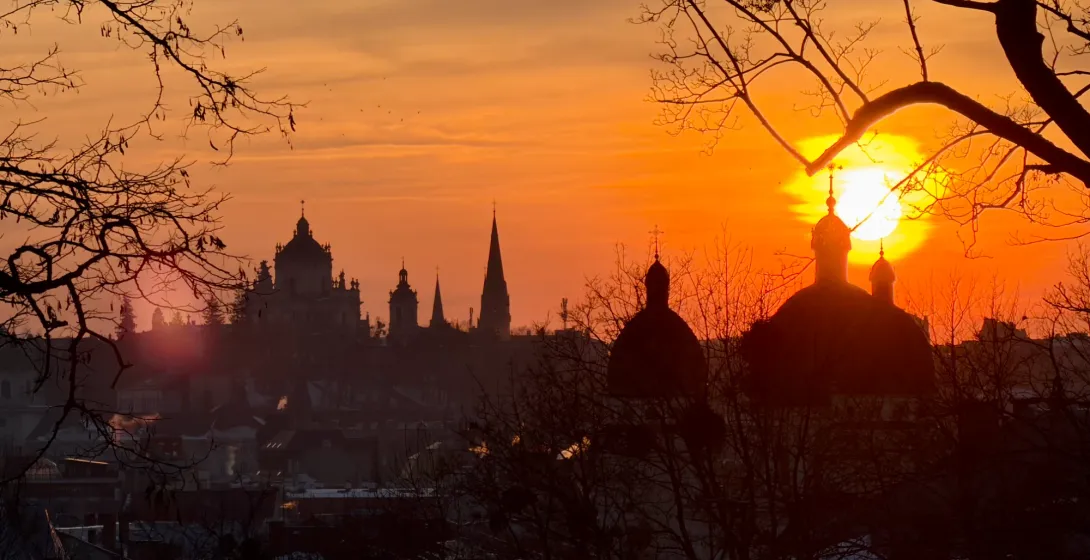Silhouettes of church domes and spires against a bright orange sunset sky, framed by bare tree branches in Lviv.