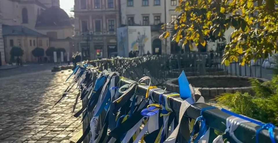 A fence along a cobblestone street in Lviv, adorned with many blue and yellow ribbons. St. Andrew's Church and historic bu...