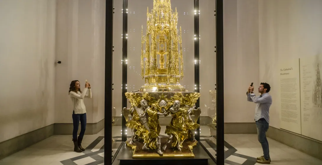Una custodia ornamentada y dorada en una vitrina de cristal, fotografiada por dos visitantes con sus teléfonos.